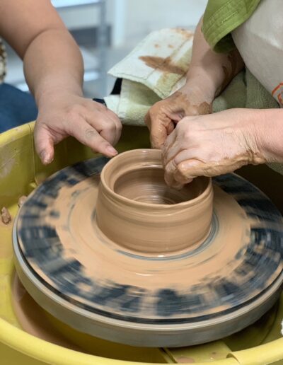 Close up of Teaching Center student hands throwing cylinder with teacher hand guiding, beginning wheel class