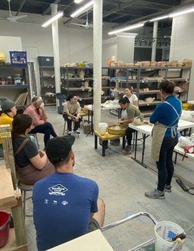 Katie Meili Messersmith demonstrating throwing off the mound at the potters wheel, surrounded by students, intermediate wheel class